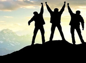 A silhouette of of 3 male figures on a mountaintop, fists raised in the air triumphantly.