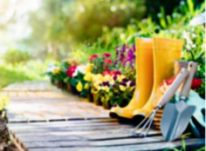 A photo of a wooden walkway through a garden. Yellow boots and garden tools rest on the ground to to the right, while colorful flowers bloom in the background.